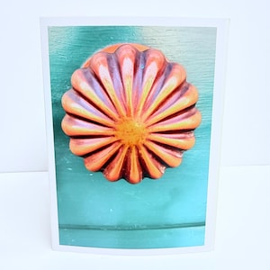 Puede incluir: Un pomo de puerta redondo y estriado con un acabado degradado en naranja y amarillo, que presenta un patr&oacute;n de rayos de sol con crestas elevadas. El pomo est&aacute; montado sobre una superficie de madera plana de color verde azulado.