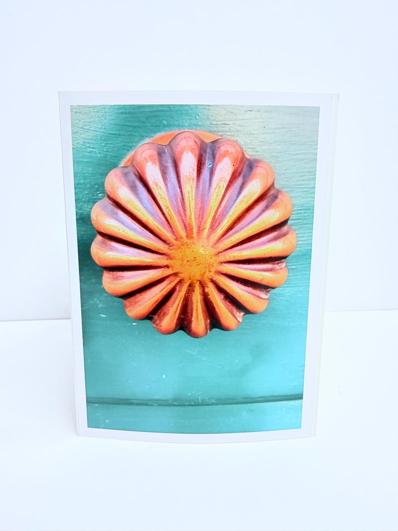 Puede incluir: Un pomo de puerta redondo y estriado con un acabado degradado en naranja y amarillo, que presenta un patr&oacute;n de rayos de sol con crestas elevadas. El pomo est&aacute; montado sobre una superficie de madera plana de color verde azulado.