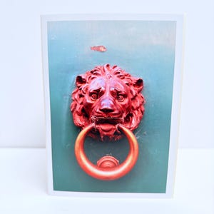 Puede incluir: Una tarjeta de felicitaci&oacute;n rectangular con una fotograf&iacute;a de un llamador de puerta en forma de cabeza de le&oacute;n de color rojo brillante con un anillo circular, montado sobre una superficie de color verde azulado mate y lisa.