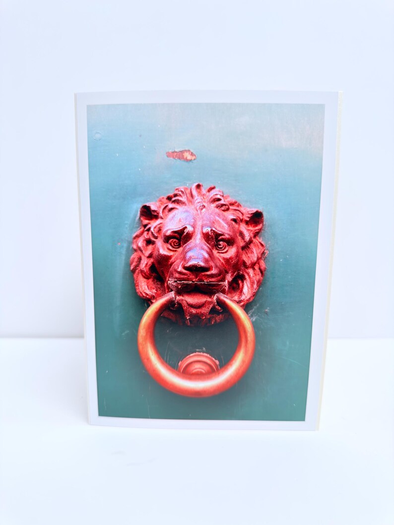 Puede incluir: Una tarjeta de felicitaci&oacute;n rectangular con una fotograf&iacute;a de un llamador de puerta en forma de cabeza de le&oacute;n de color rojo brillante con un anillo circular, montado sobre una superficie de color verde azulado mate y lisa.