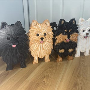 Peut inclure: Quatre figurines de chiens en bois représentant des Poméraniens. Les figurines sont peintes en noir uni, beige clair, noir et beige, et blanc. Chaque chien présente une texture de fourrure sculptée, des yeux ronds, un nez noir et une langue rose.