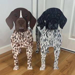 May include: Two freestanding wooden dog sculptures shaped like German Shorthaired Pointers. One figure features a brown head with a white blaze and a brown and white spotted coat. The other figure has a black head and a black and white spotted coat.