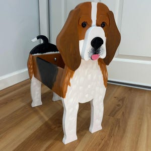 May include: A wooden planter shaped like a beagle dog with brown, white, and black painted details. The dog figure features long brown ears, a white muzzle, a black nose, and a pink tongue. The hollow body serves as a container for plants.