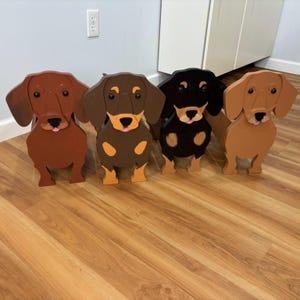 May include: Four wooden dachshund dog figures in various shades of brown and black. Each figure has a distinct color scheme, with details like ears, paws, and faces painted in contrasting colors. The dogs are standing upright.