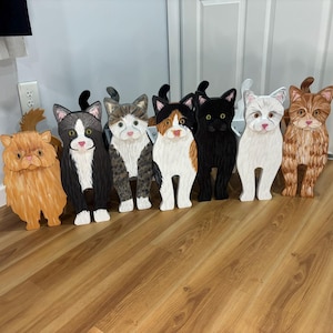 May include: A collection of seven standing wooden cat figurines featuring various coat patterns including orange long-haired, black and white, grey tabby, calico, solid black, white, and orange tabby, all displayed in a row on a wooden floor.