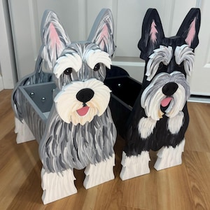 May include: Two wooden dog-shaped storage bins featuring Schnauzer designs. One bin is painted in shades of gray and white, while the other is black and white. Both feature carved details, pointed ears, white eyebrows, and open mouths with pink tongues.