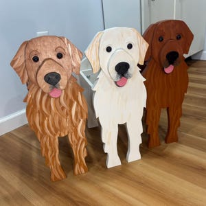 May include: Three wooden dog sculptures in varying shades of brown and white. Each dog has a distinct color, with painted details including eyes, noses, and pink tongues. The sculptures are freestanding and appear to be decorative.