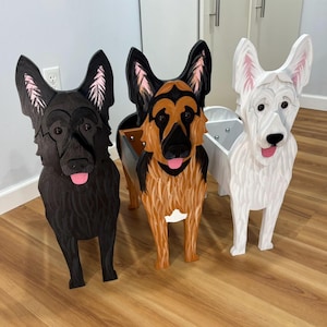 May include: Three wooden German Shepherd dog planters. One is black, one is brown and tan, and the other is white. Each has a pink tongue and inner ear detail. The planters are standing on a wood floor.
