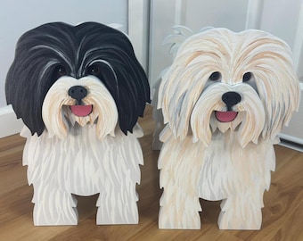 May include: Two standing dog figurines crafted from wood. The left figurine features black and white fur with a black face and white muzzle. The right figurine features cream and white fur. Both have black noses, dark eyes, and pink tongues.