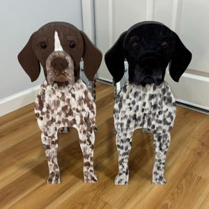 May include: Two wooden dog sculptures. One is brown and white, the other is black and white. Both have a spotted pattern on their bodies and are standing on four legs. The sculptures are detailed with realistic eyes and facial features.
