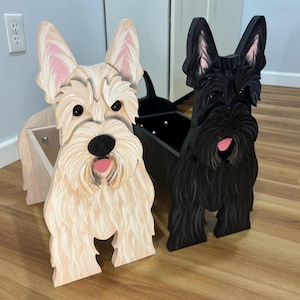 May include: Two wooden dog-shaped planters. One is painted light beige with pink accents on the ears and tongue, the other is black with pink accents. Both have open compartments for plants.