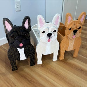 May include: Three wooden French bulldog planters in black, white, and tan. Each dog has pink-lined ears and a pink tongue sticking out. The black dog has a white chest, and the white dog has a black nose.