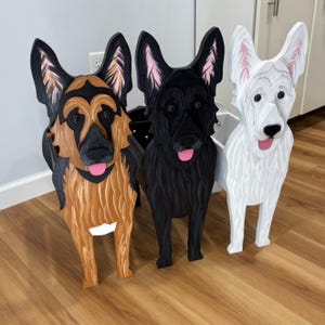 May include: Three standing dog-shaped wooden cutouts featuring German Shepherd designs. One is tan and black, one is solid black, and one is solid white. Each figure has carved fur textures, pointed ears, dark eyes, and a pink tongue visible.