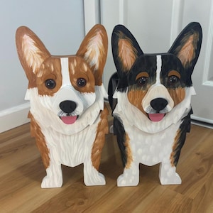 May include: Two standing wooden corgi figures, one with tan and white fur and the other with black, tan, and white fur. Both figures feature pointed ears, dark eyes, black noses, and pink tongues, with detailed brushstroke textures on their surfaces.