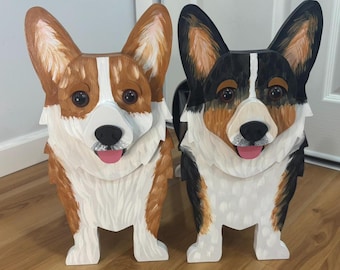 May include: Two standing wooden corgi figures, one with tan and white fur and the other with black, tan, and white fur. Both figures feature pointed ears, dark eyes, black noses, and pink tongues, with detailed brushstroke textures on their surfaces.