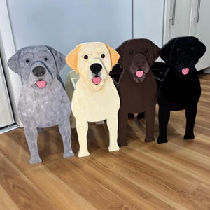 May include: Four wooden Labrador Retriever dog sculptures in grey, cream, brown, and black. Each dog has a pink tongue and is standing upright. The sculptures are painted with details to resemble the breed.