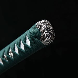 May include: Close-up of a katana handle. The handle is wrapped in dark green fabric with white diamond-shaped accents. The end cap is silver-colored with an ornate, swirling design. The background is black.