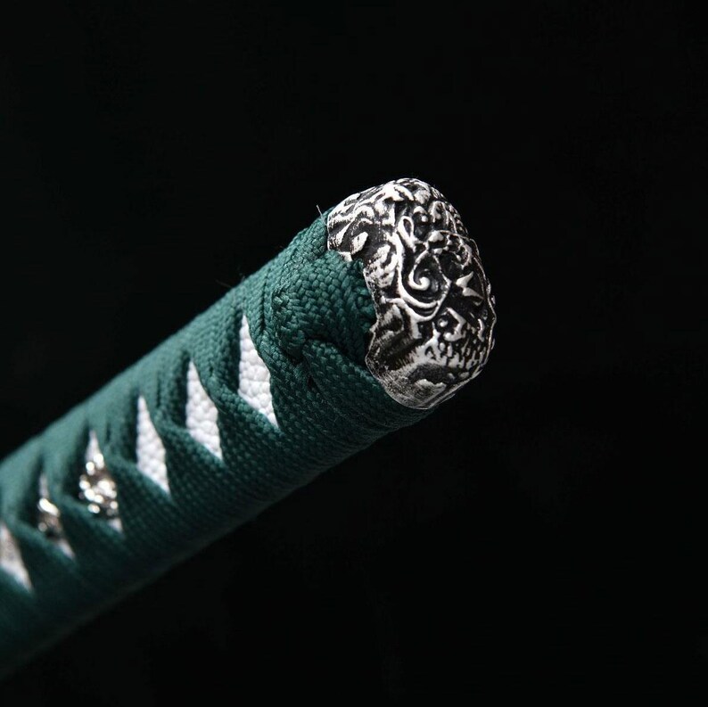 May include: Close-up of a katana handle. The handle is wrapped in dark green fabric with white diamond-shaped accents. The end cap is silver-colored with an ornate, swirling design. The background is black.