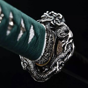 May include: Close-up of a katana handle with a detailed silver dragon design. The handle features a dark green wrap with white accents. The dragon's scales and features are intricately carved, showcasing a blend of artistry and craftsmanship.