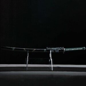 May include: A Japanese katana sword with a curved black blade and a green handle. The blade has a decorative pattern, and the handle is wrapped in a diamond pattern. The sword is displayed on a clear acrylic stand against a black background.