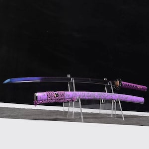 Op de afbeelding: Een katana-zwaard met een paarse handgreep en schede, en een zwart lemmet met blauwe accenten. De schede heeft een textuurpatroon. Het zwaard wordt op een transparante standaard tegen een donkere achtergrond getoond.