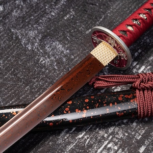 May include: A Japanese katana sword with a red handle and a brown blade featuring a unique pattern. The sword's guard is silver and gold, and the scabbard is black with red accents. The blade is approximately 18 inches long.