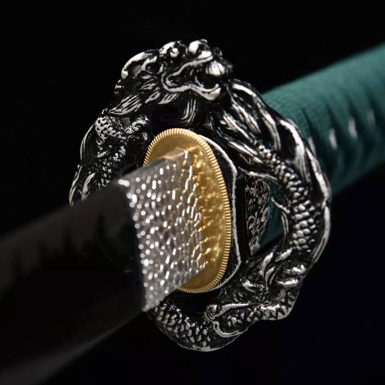 May include: Close-up of a katana sword with a detailed dragon-shaped guard. The guard is silver with intricate detailing, surrounding a gold-colored disc. The blade is black, and the handle is wrapped in green fabric.