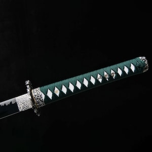 May include: A detailed view of a katana sword. The handle is wrapped in green fabric with white diamond-shaped accents. The blade is black and silver, with intricate metalwork near the guard. The background is a solid black.