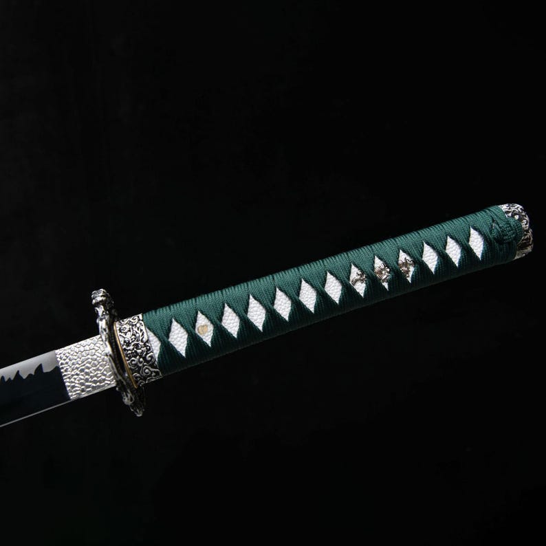 May include: A detailed view of a katana sword. The handle is wrapped in green fabric with white diamond-shaped accents. The blade is black and silver, with intricate metalwork near the guard. The background is a solid black.
