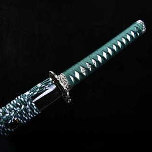May include: A close-up of a katana sword with a green handle and a silver guard. The handle is wrapped in green fabric with white diamond-shaped accents. The blade is black and the guard has an ornate design.