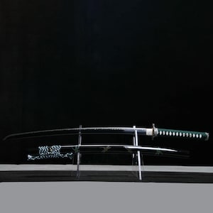 May include: A Japanese katana sword with a curved, black blade and a green handle with white diamond-shaped accents. The sword rests on a clear acrylic stand, with the scabbard below. The background is a solid black.