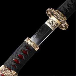 May include: Close-up of a katana sword. The handle is wrapped in black cord with red accents. The blade is black with etched designs. Gold-colored metal accents with dragon motifs adorn the hilt and other parts of the sword.