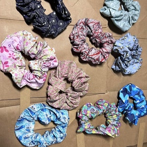 May include: A collection of ten colorful fabric scrunchies with various patterns, including floral, geometric, and abstract designs. The scrunchies are arranged on a brown paper background.