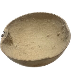 May include: A beige, oval-shaped bowl with a rough, textured surface. The bowl has a natural, organic appearance, with a slightly uneven rim and visible imperfections. The interior of the bowl is a lighter shade of beige.