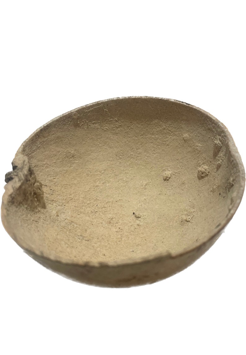 May include: A beige, oval-shaped bowl with a rough, textured surface. The bowl has a natural, organic appearance, with a slightly uneven rim and visible imperfections. The interior of the bowl is a lighter shade of beige.