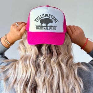 May include: A pink trucker hat with a white mesh back. The front of the hat features a black and white graphic of a bison with the text "Yellowstone National Park".