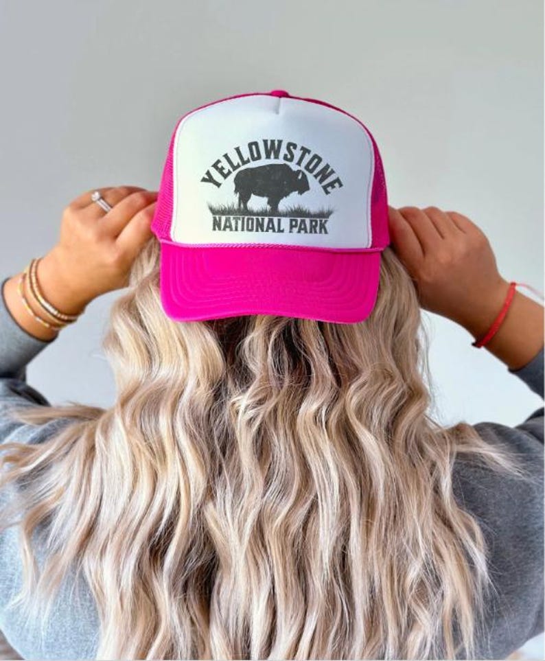 May include: A pink trucker hat with a white mesh back. The front of the hat features a black and white graphic of a bison with the text "Yellowstone National Park".