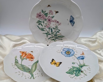 May include: Three white ceramic plates with scalloped edges feature floral and butterfly designs. The plates have a delicate, feminine aesthetic.