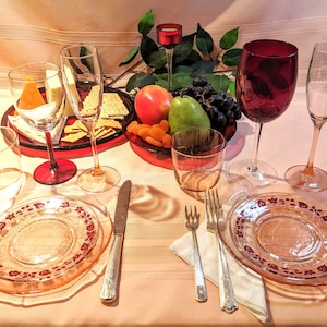 May include: A table setting with two pink glass plates with a floral design, two silver forks, two silver knives, two clear glasses, a red wine glass, a clear champagne glass, a red glass with a candle, a plate of crackers, a plate of fruit, and a plate of dried apricots.