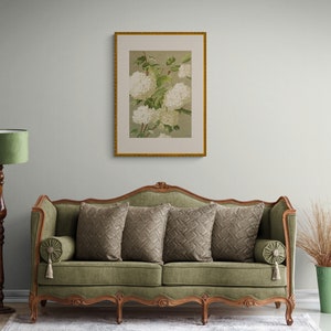 May include: A green velvet sofa with a carved wooden frame and four legs. The sofa has three pillows with a geometric pattern and a tasseled armrest. A framed print of white flowers hangs on the wall behind the sofa.
