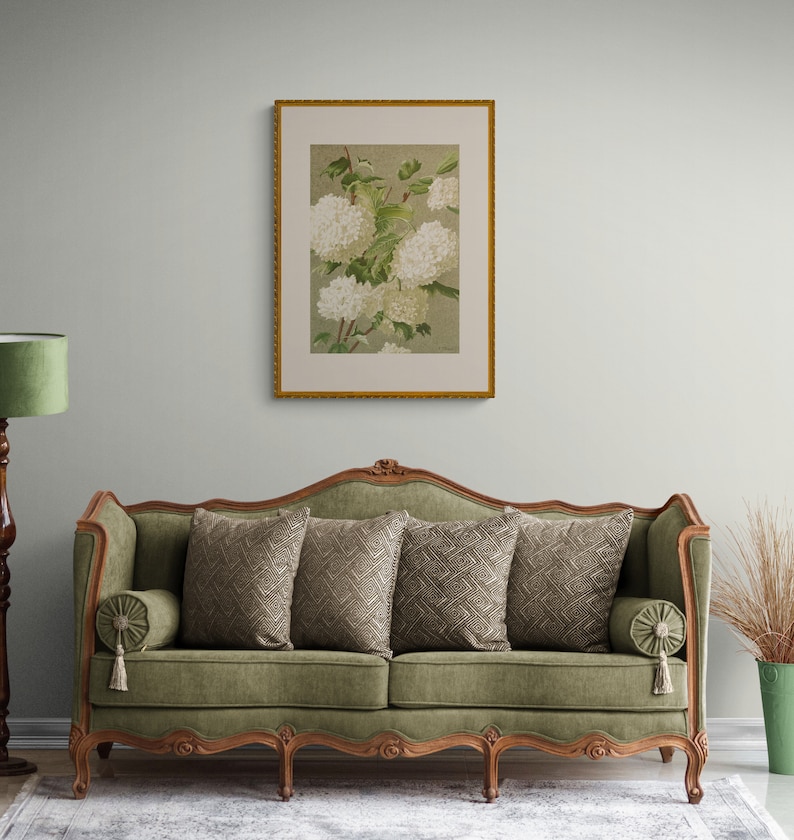 May include: A green velvet sofa with a carved wooden frame and four legs. The sofa has three pillows with a geometric pattern and a tasseled armrest. A framed print of white flowers hangs on the wall behind the sofa.