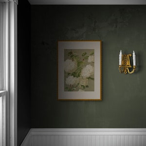 May include: A framed botanical print of white flowers with green leaves hangs on a dark green wall. A gold wall sconce with two candles is on the right side of the frame.