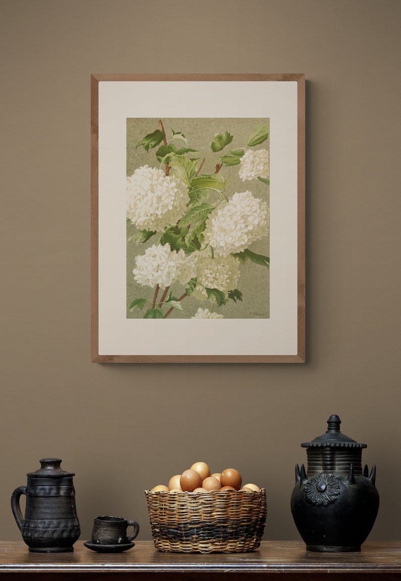 May include: A framed botanical illustration of white flowers with green leaves. The image is in a wooden frame and is hanging on a brown wall. There are two black ceramic jars and a basket of eggs on a wooden table in the foreground.