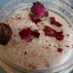 May include: A clear glass jar filled with white bath salts and dried rose petals and buds. The bath salts have a coarse texture and are interspersed with small pink granules. The rose petals are a deep red color.