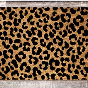 Puede incluir: Una alfombra rectangular con un estampado de leopardo. La alfombra presenta un fondo marrón claro con manchas negras, creando un diseño audaz y elegante. La alfombra probablemente está hecha de un material duradero adecuado para uso en exteriores.