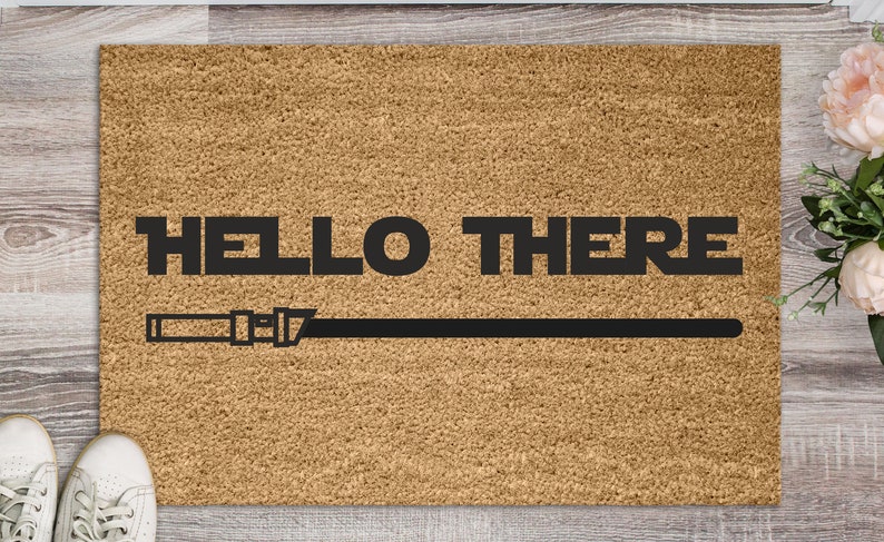 May include: A brown coir doormat with the text "HELLO THERE" in black lettering and a black lightsaber graphic.