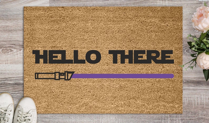 May include: A brown coir doormat with the text "HELLO THERE" in black lettering and a purple lightsaber graphic.