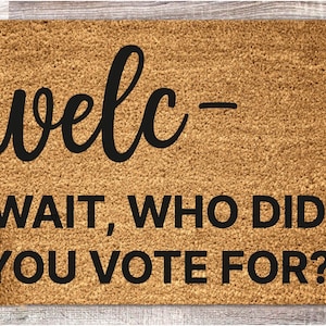 May include: A rectangular doormat with a light brown, textured surface. The mat features the word "welc-" in a black, cursive font, followed by the phrase "WAIT, WHO DID YOU VOTE FOR?" in a bold, sans-serif font.