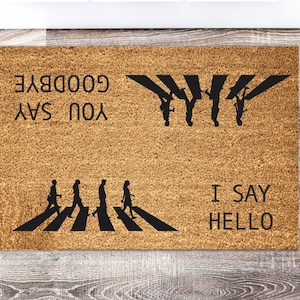 Op de afbeelding: Een rechthoekige deurmat van natuurlijke kokosvezel. De mat heeft een zwarte afbeelding van mensen die een zebrapad oversteken, met de tekst "YOU SAY GOODBYE" en "I SAY HELLO" in zwarte letters. Een decoratief item voor in huis.
