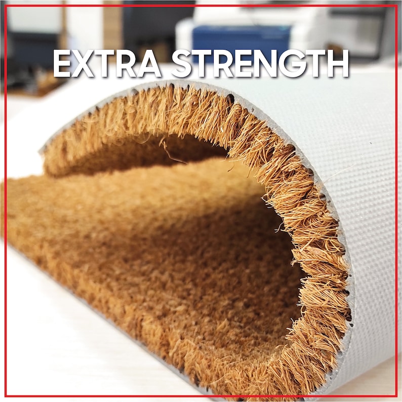 May include: Close-up of a brown coir doormat with the text "EXTRA STRENGTH" printed on the top. The mat is folded in half, revealing the backing material.